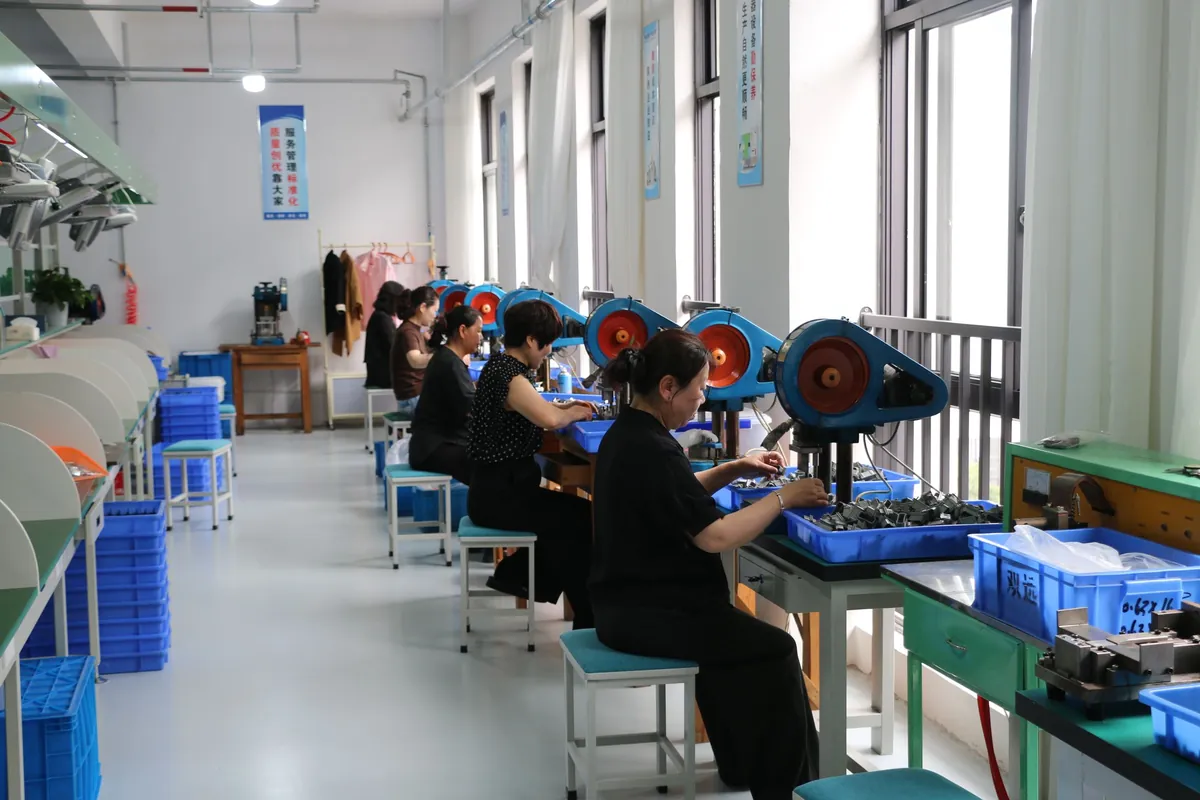 Operators at workstations along the window wall, feeding small parts from blue bins